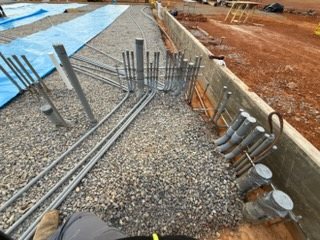 Conduit stub-ups, bends, and offsets laid in gravel beside a foundation wall before a slab pour.